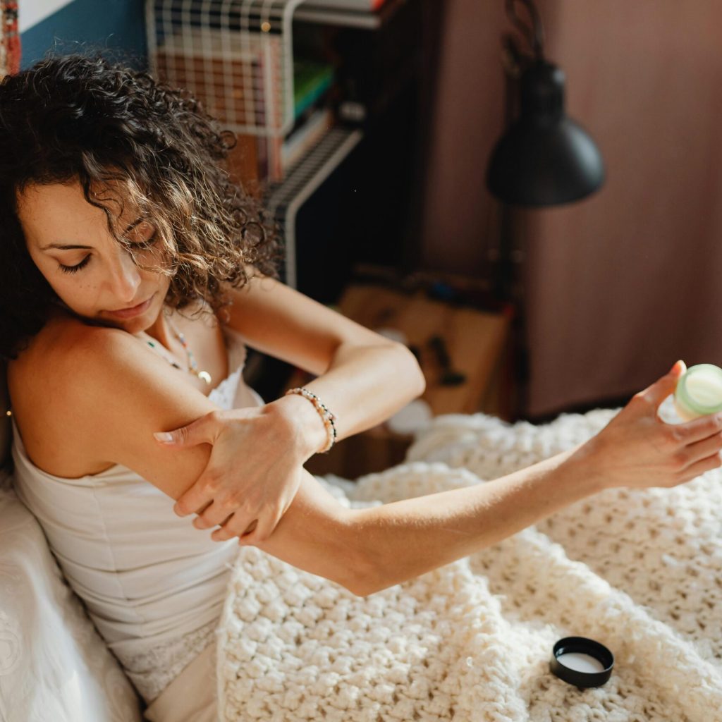 Woman applying skincare lotion in a cozy bedroom, focusing on relaxation and self-care.