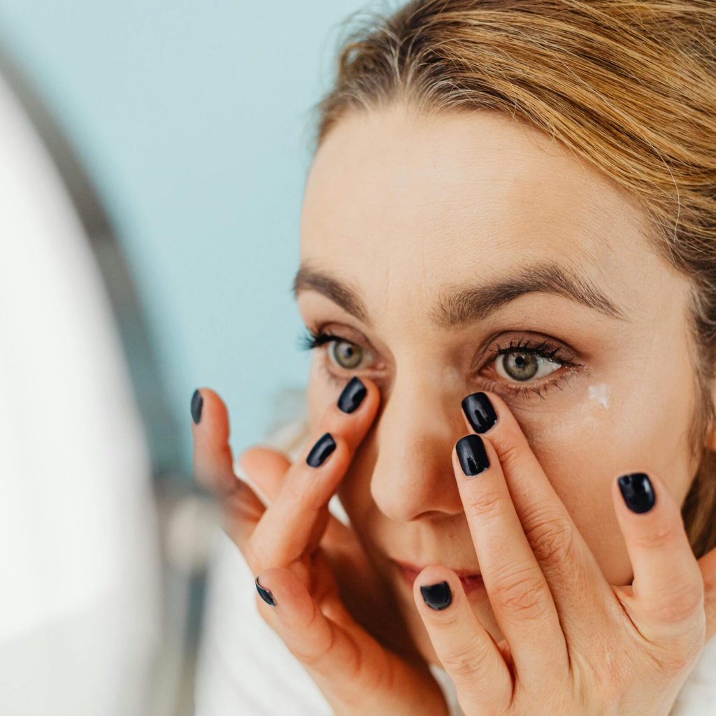 Close-up of a woman applying facial cream in front of a mirror with blue background.